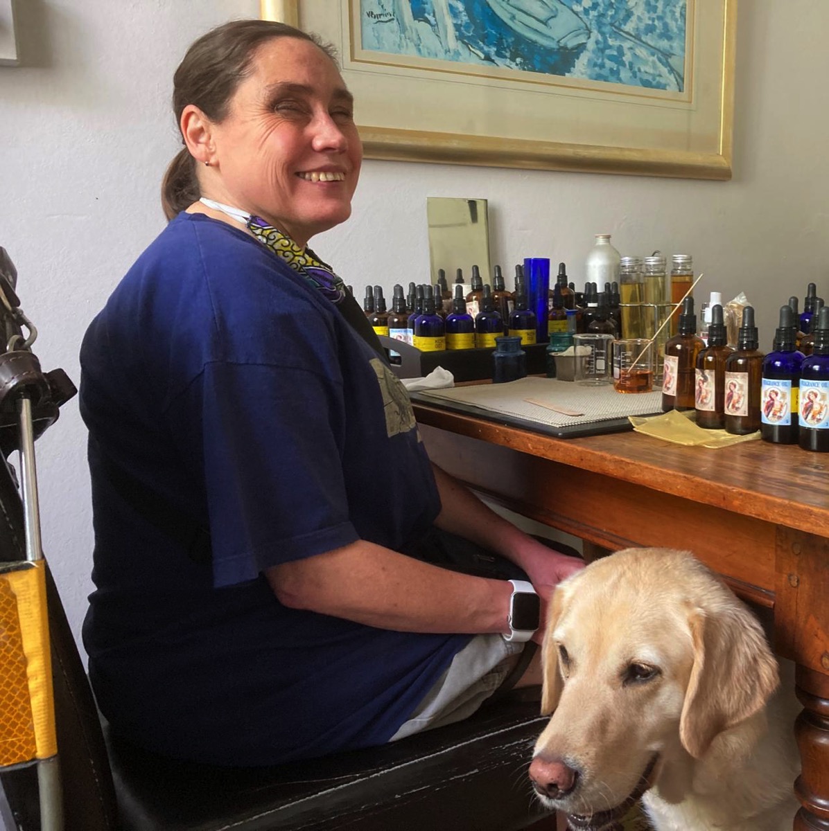 The image shows Lois sitting at a table surrounded by fragrance bottles and Fiji standing beside her.