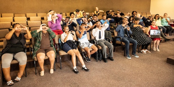 the image shows a group of people posing for the photograph,, holding their hands up to their face making a square through which they are looking. In the front row a laptop rests on the lap of one woman, showing another woman’s face on the screen. 