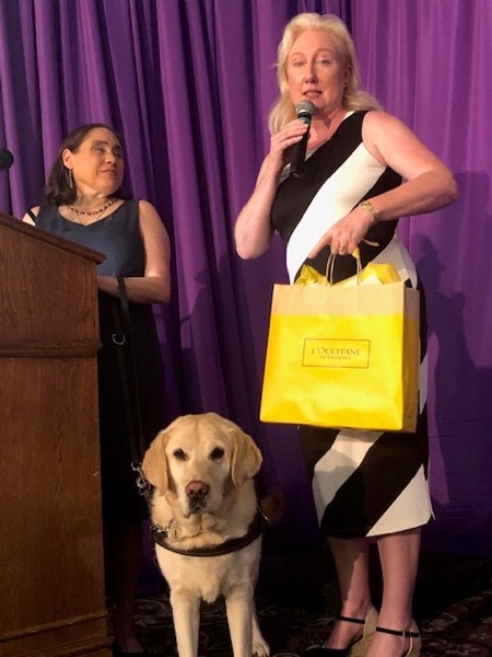 he image shows two women standing on a stage behind a lectern, with a Yellow Labrador standing next to them. The woman on the right is handing a gift bag to the woman on the left.