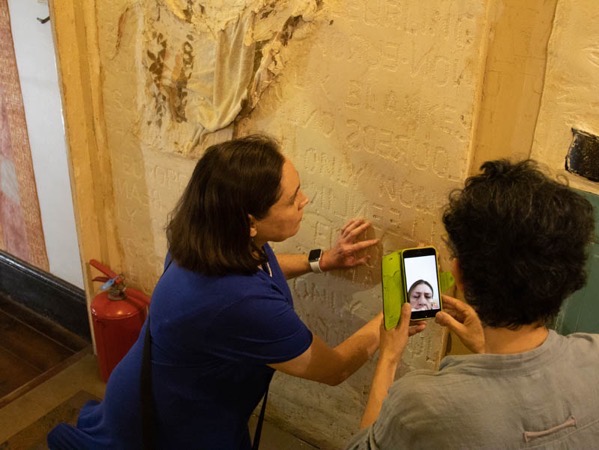 A blind woman with shoulder-length dark hair kneels in front of a wall that is part of a museum installation and touches the writing engraved in its plasterwork. She wears a short-sleeved blue dress. To her right, is the shoulder and head of a woman with short dark hair wearing a grey top, who has her back to the viewer holding a mobile phone. A third woman’s face can be seen on the phone screen, observing the exploration via WhatsApp.