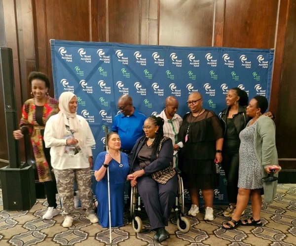 Image of a group of 9 people in front of a banner reading Road Accident Fund.  The people are of diverse genders and races, with one woman being a wheelchair user and another woman being a white cane user indicating she is blind.