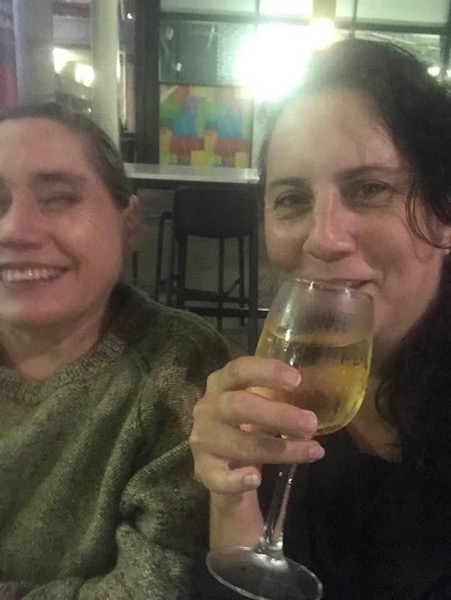 Two women leaning close and smiling, toasting with glasses of wine. The woman on the left has long dark hair and is wearing a light green sweater. The woman on the right has light brown hair. They are pictured in a restaurant. 
