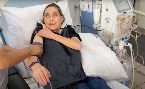 A woman with dark hair that is pulled back is lying on a reclinable hospital bed. She is wearing a dark blue sleeveless fleece top and is wearing a blood pressure cuff. Behind her a dialysis machine is visible
