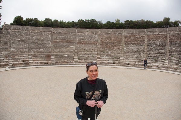 A woman standing in the center of an ancient stone amphitheatre in Greece. She has brown hair pulled back, and is wearing a black sweatshirt with a design on the front, sunglasses on her head, and a crossbody bag. She is smiling and holding a white cane in her hands. The amphitheatre has many rows of stone seating that curve around in a semi-circle, and there is another person walking in the background near the seats. The sky above is cloudy, and there are trees visible at the top of the amphitheatre.
