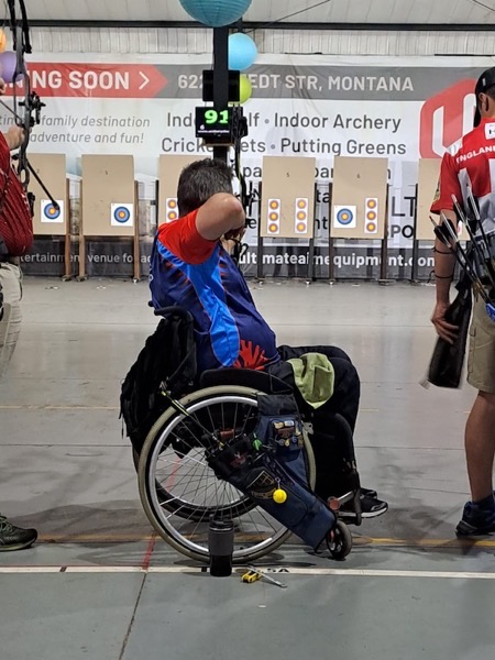 Ferdi, A man in a wheelchair, who is wearing a blue and red shirt is aiming a bow, presumably at  an archery target, On the wheelchair there is a dark blue bag containing various articles. Other competitors can be seen in the background as are archery targets and a digital scoreboard.