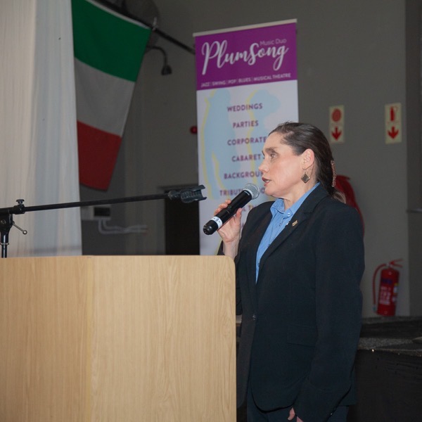 Woman with dark hair wearing a dark suit and blue top standing behind a wooden podium, holding a microphone and speaking. On the left, a large flag with green, white, and red stripes is hanging. The setting is indoors at an event.