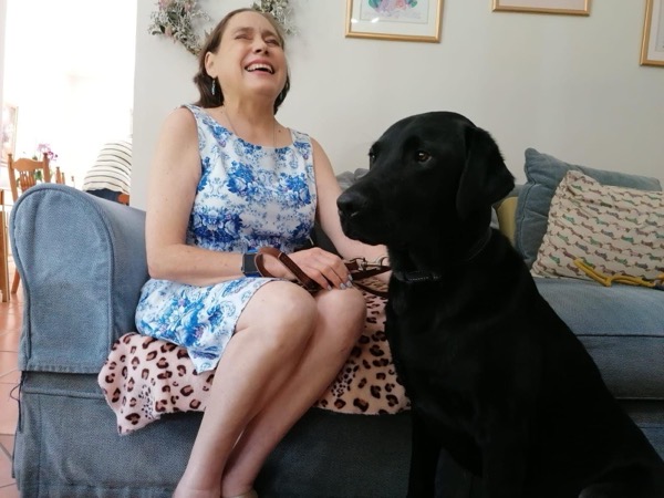A dark-haired woman sitting on a blue couch, smiling, with a black dog at her side. The woman is wearing a sleeveless blue and white floral dress and has a watch on her left wrist. She is holding the guide dog’s harness. A pink blanket with brown spots is draped over the couch under her. The dog is large, sitting upright and looking forward. Behind them, there are decorative pillows, one with a pattern of dachshunds, and a wall with framed pictures and a floral wreath. There’s also a glimpse of a dining area in the background, with someone sitting at a table. The overall scene is warm and homely.